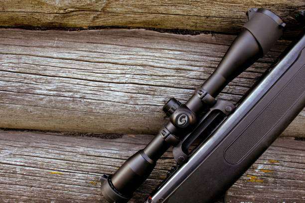 Part of a black rifle with a telescopic sight on a wooden background.